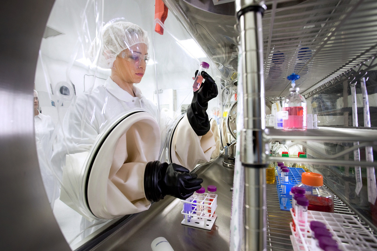 Lab worker in PPE handles vials inside a sealed isolator, performing Biologics testing with samples and instruments in a controlled lab environment