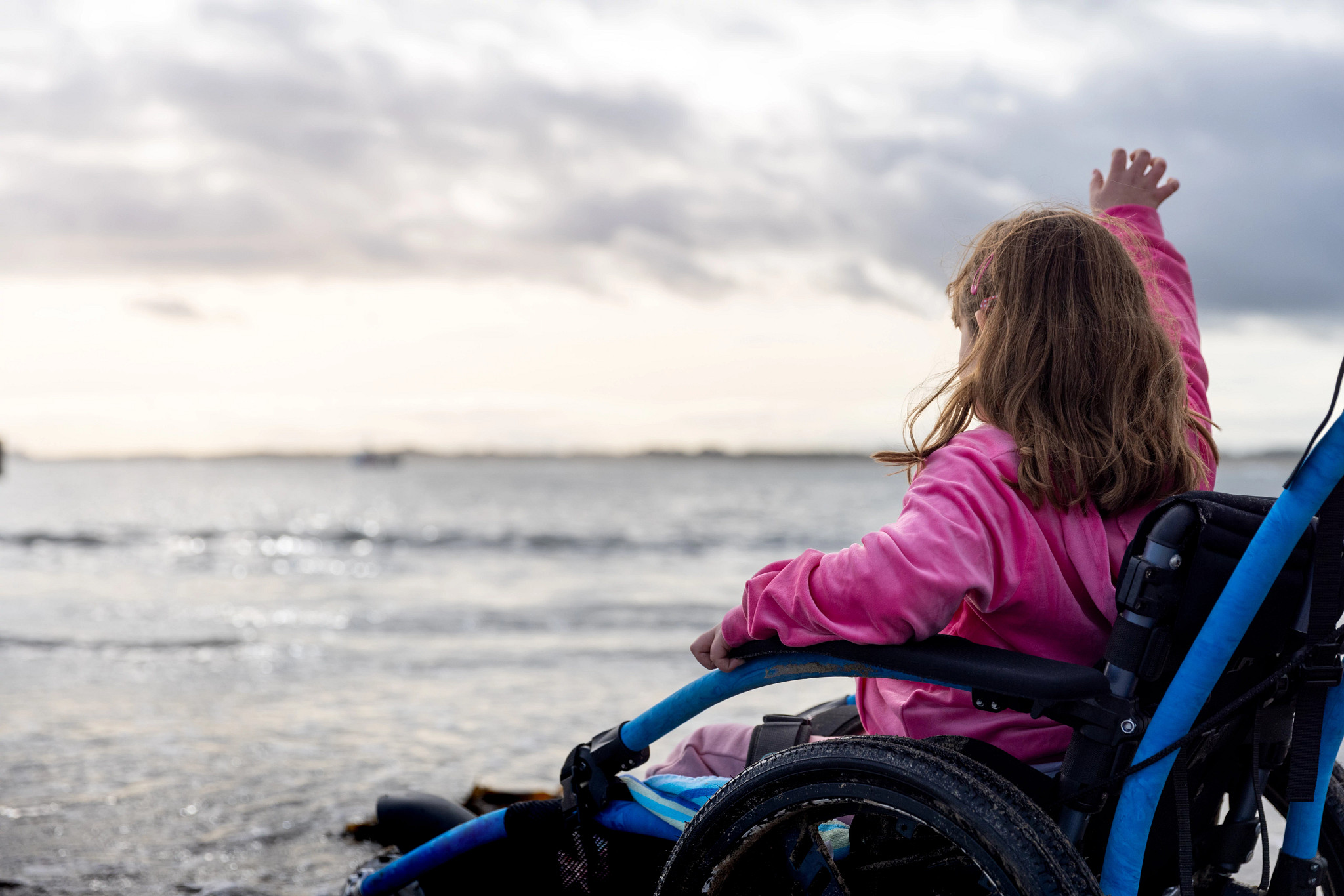 Photo of a little girl in a wheelchair. She is wearing pink and looking out over the ocean while waving.