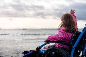 Photo of a little girl in a wheelchair. She is wearing pink and looking out over the ocean while waving.