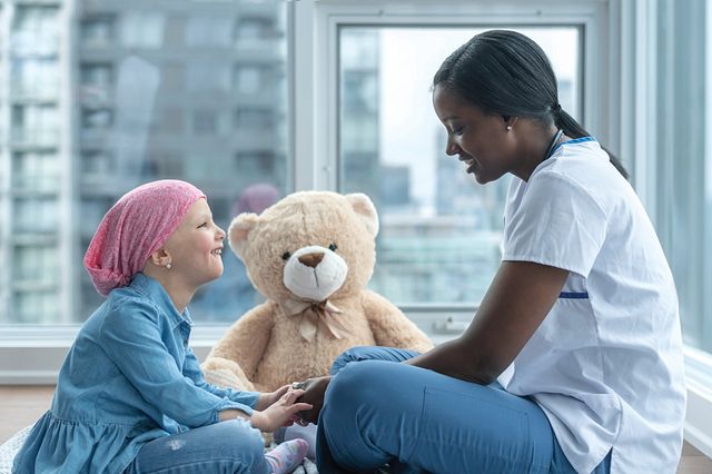 Nurse sitting with a child cancer patient and smiling. Teddy bear sits in the background.