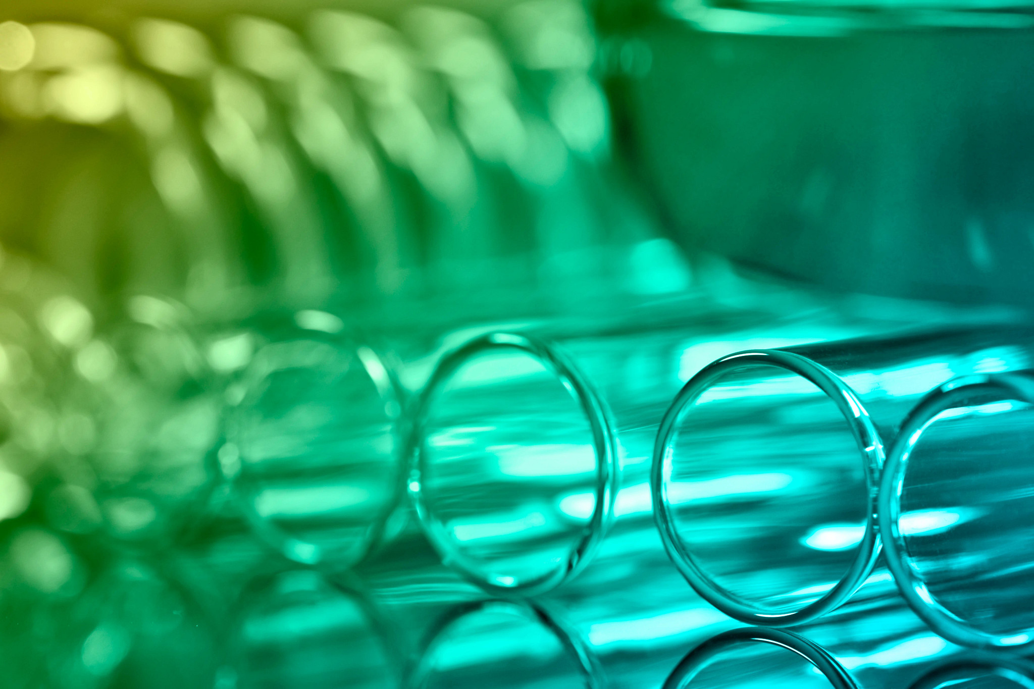 Close-up of clear glass test tubes arranged horizontally on a reflective surface, with green and teal lighting and a blurred laboratory-style background.