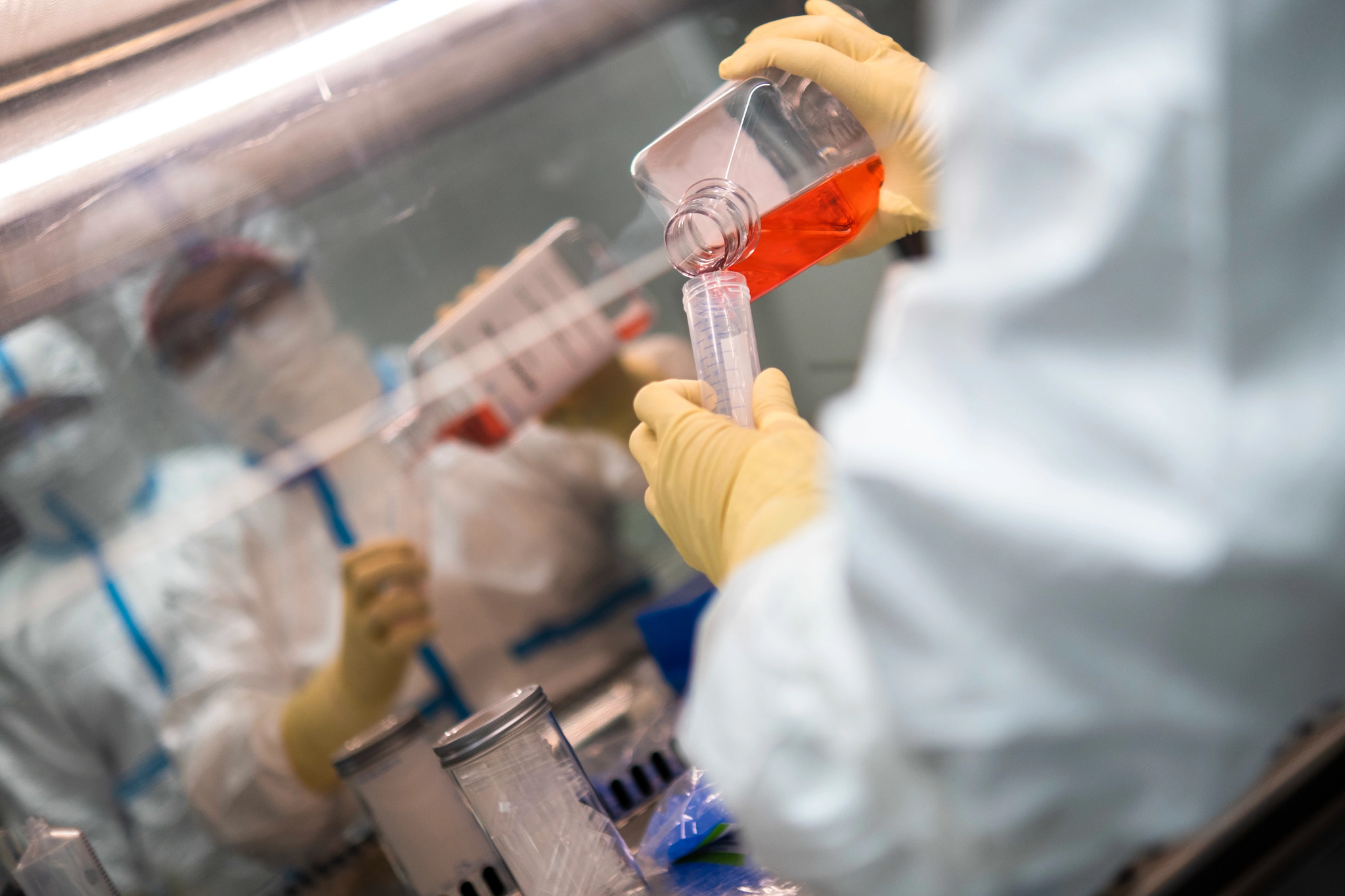 Scientists hands pouring an orange liquid from a larger bottle into a test tube.
