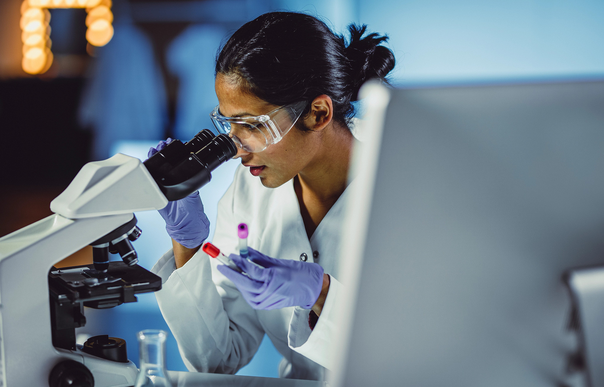 Female scientist wearing PPE and looking through a microscope with two vials in her hand.