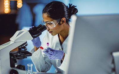 Female scientist wearing PPE and looking through a microscope with two vials in her hand.