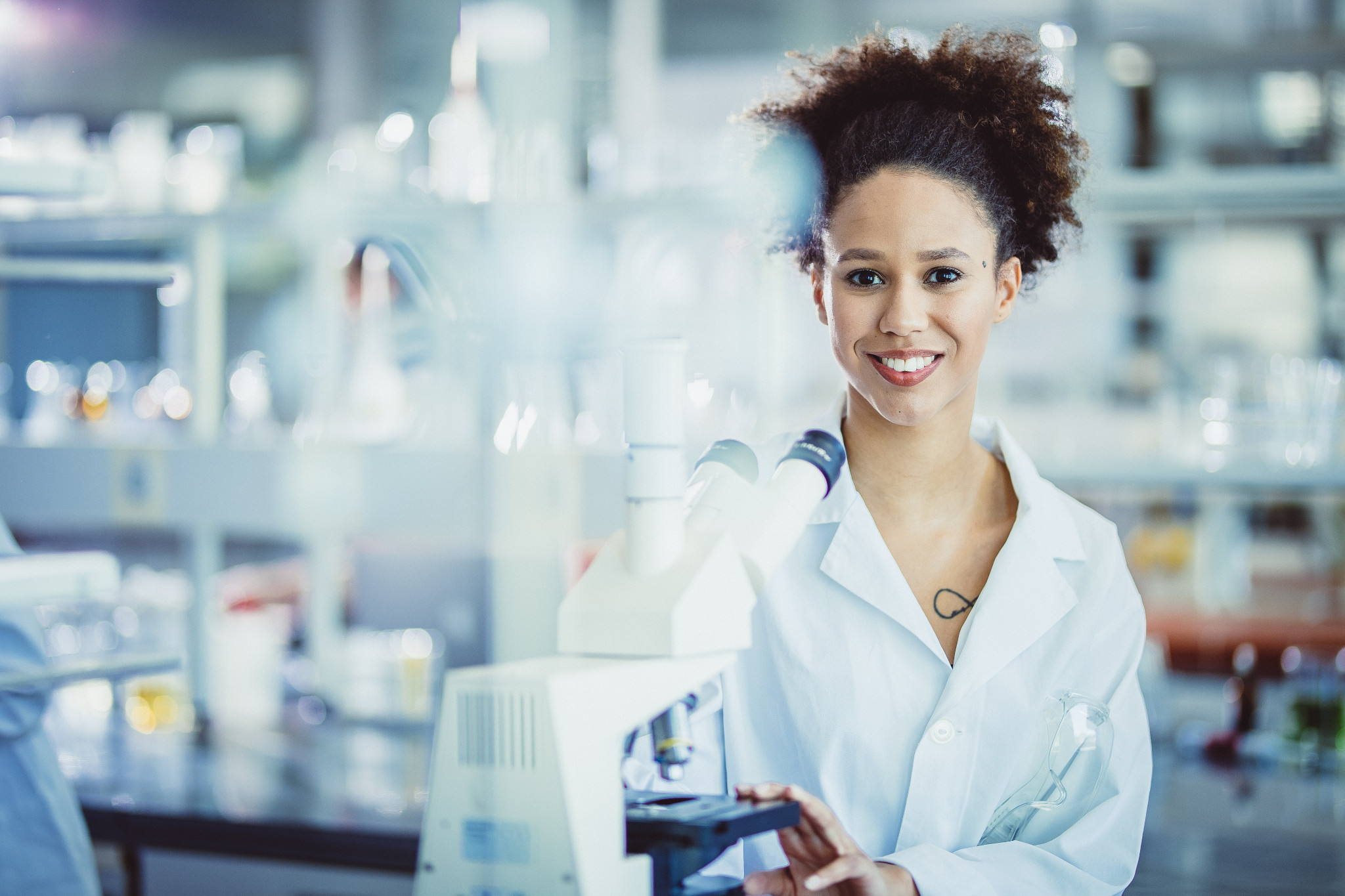 Female scientist with curly hair and wearing a lab coat standing in a lab next to a microscope.