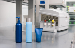 Cosmetic bottles and tube on a lab bench with microbiology testing equipment in the background, representing quality control in home and personal care manufacturing