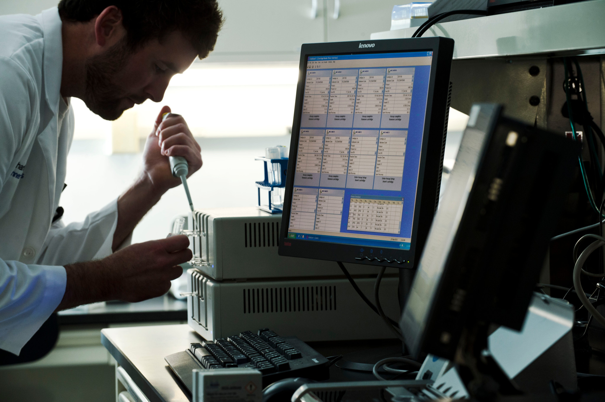 Image of a man in a lab coat, holding a pipette, dispensing sample into a cartridge on a laboratory instrument.