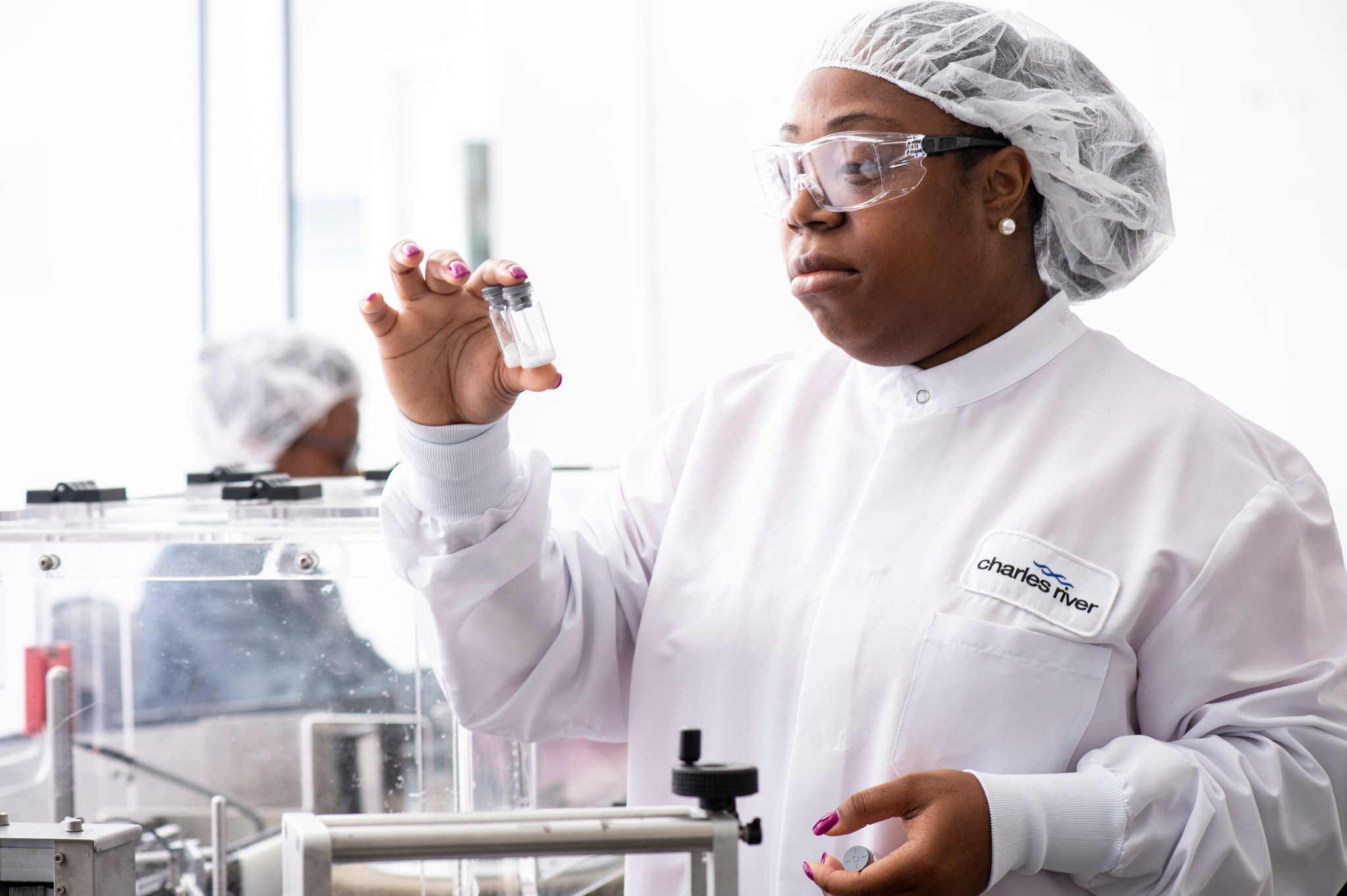 Image of a woman in a lab coat wearing safety glasses, holding two vials together analyzing them.