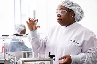Image of a woman in a lab coat wearing safety glasses, holding two vials together analyzing them.