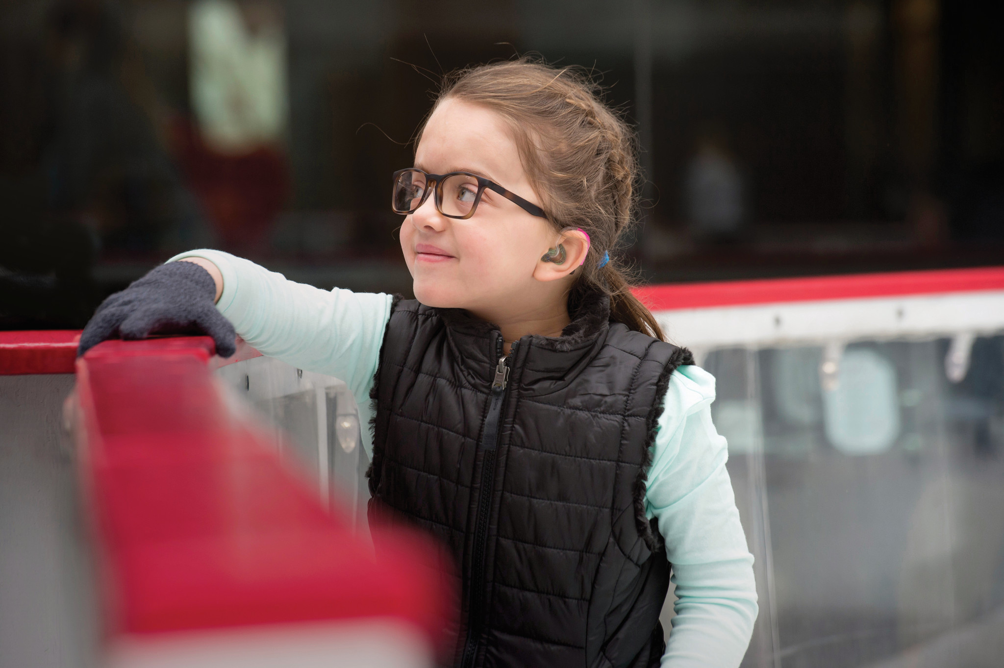 Photo of a young girl named Evie, wearing glasses and gloves, looking away from the camera at an ice skating rink.