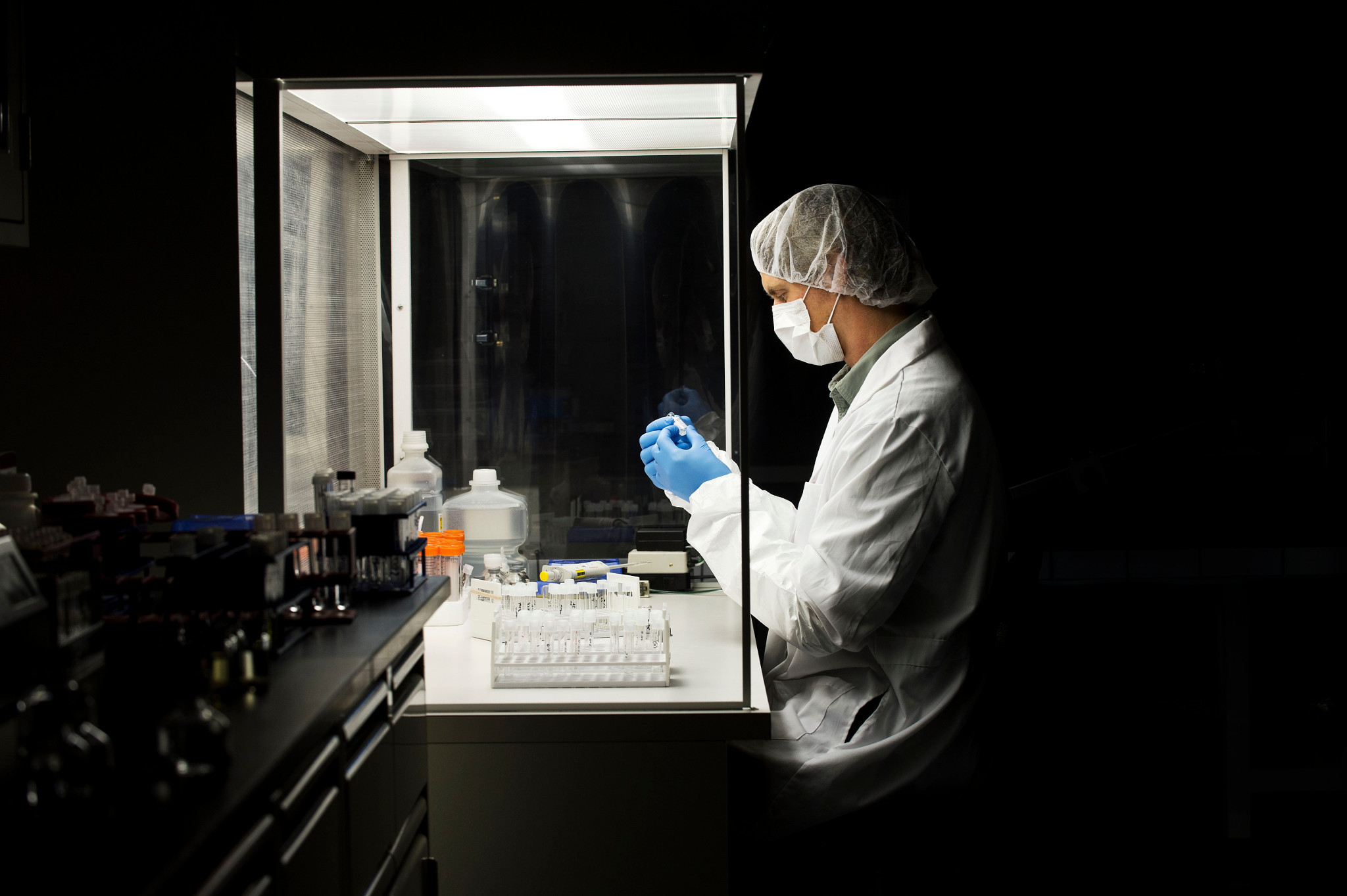 Image of a man wearing a lab coat working in a hood, looking at a test tube, surrounded by laboratory equipment and materials.