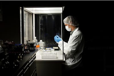 Image of a man wearing a lab coat working in a hood, looking at a test tube, surrounded by laboratory equipment and materials.