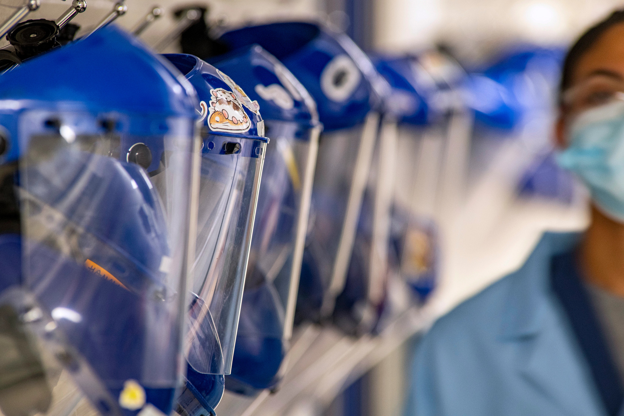 Row of protective face shields and respirator helmets used in a toxicology research lab, with a scientist blurred in the background.