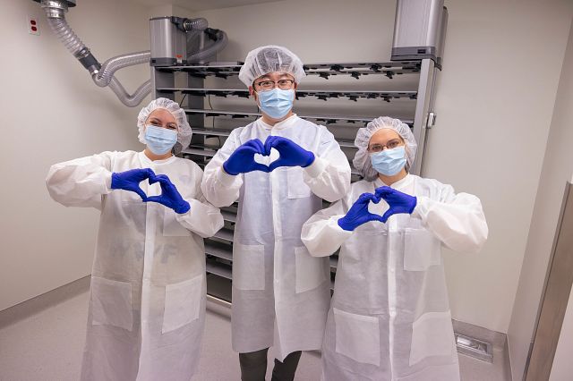 three lab techs in white coats, hair nets, and blue gloves making a heart with their hands